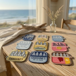 Può includere: Una collezione di nove sottobicchieri a tema sardine in vari colori, tra cui blu, giallo e rosa. Ogni sottobicchiere presenta un design di scatola di sardine con la scritta "Sardines" o "Sardines de la Mer". I sottobicchieri sono disposti su un tavolo di legno.