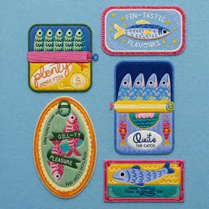 May include: Embroidered patches with fish themes. Designs include sardines in cans, a fish with the text "Fin-tastic Flavours", and other fish-related phrases. The patches are on a light blue background.