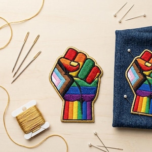 Op de afbeelding: Geborduurde patch in de vorm van een vuist, met een regenboogontwerp en gouden rand. De patch is geplaatst op een denim stof. Naainaalden, spelden en gouden draad zijn rond de patch gerangschikt.
