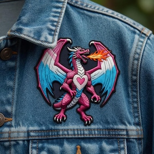 Könnte beinhalten: Gestickter Aufnäher mit einem rosa Drachen mit blau-weißen Flügeln, der Feuer speit. Der Drache hat eine herzförmige Brust. Der Aufnäher ist auf einer Jeansjacke angebracht.