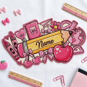 Pode incluir: Um emblema bordado com tema escolar apresentando um lápis amarelo com a palavra Name em cursivo, rodeado por ícones cor-de-rosa, incluindo um caderno, maçã, globo, lápis de cor e laços. Uma régua de madeira mostra 23 cm de comprimento.
