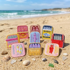 May include: A collection of nine rectangular embroidered patches featuring various sardine and anchovy tin designs. The patches display colorful illustrations of fish, text including SARDINAS, SARDINES, and ANCHOVIES, and metallic-look pull tabs, all arranged on a sandy beach.