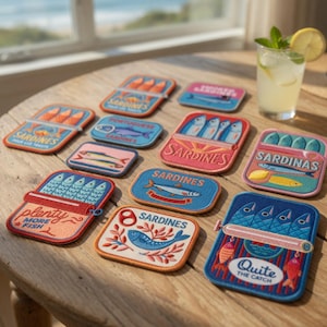 Può includere: Una collezione di toppe colorate a tema sardine su un tavolo di legno. Le toppe presentano vari disegni, tra cui scatole di sardine aperte, illustrazioni di pesci e testo come "Sardines" e "Quite the Catch". È visibile anche una bevanda rinfrescante.