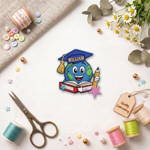 May include: Embroidered patch featuring a smiling globe wearing a graduation cap with the name "WILLIAM". The globe holds a pencil and sits atop an open book. The patch is surrounded by sewing supplies and decorative elements.