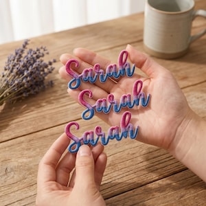 Puede incluir: Tres parches bordados con el nombre "Sarah" en letra cursiva. Los parches presentan un degradado de hilo rosa y azul con un contorno blanco. Los parches están sostenidos en las manos de una persona.