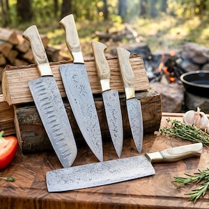 Op de afbeelding: Een set van zes keukenmessen met licht houten handvatten en stalen lemmeten met patronen. De messen zijn gerangschikt op een houten snijplank, met een natuurlijke buitenachtergrond. De messen hebben een decoratief patroon en zijn van verschillende groottes.