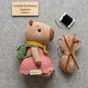 May include: Crocheted capybara doll wearing a pink dress, green backpack, and yellow scarf. The doll has a light brown body, a brown snout, and black eyes. A wooden sign reads "crochet by hamza Capybara Pattern". Also shown are yarn, crochet hooks, and black thread.