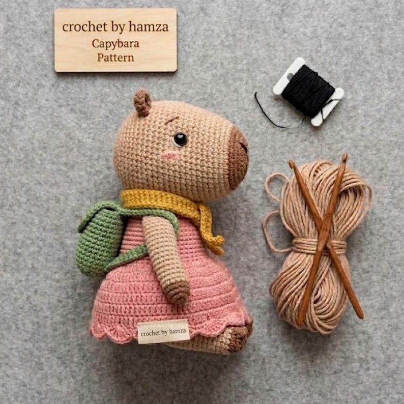 May include: Crocheted capybara doll wearing a pink dress, green backpack, and yellow scarf. The doll has a light brown body, a brown snout, and black eyes. A wooden sign reads "crochet by hamza Capybara Pattern". Also shown are yarn, crochet hooks, and black thread.