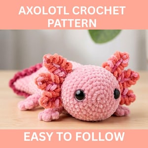 May include: A pink crocheted axolotl with black safety eyes and orange-pink gills. The text "AXOLOTL CROCHET PATTERN" and "EASY TO FOLLOW" are displayed above and below the axolotl. The plush toy is on a wooden surface.