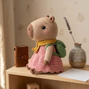 May include: A handmade crochet capybara doll wearing a pink dress, yellow scarf, and green backpack. The doll has a light brown body, dark brown nose, and black eyes. It is sitting on a wooden shelf.
