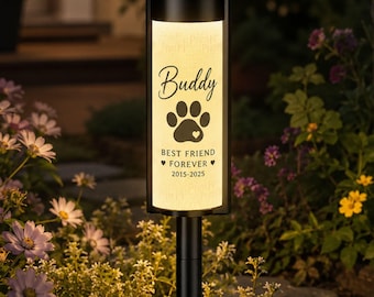 Puede incluir: Una lámpara solar negra para exteriores con un panel rectangular iluminado. El panel muestra el nombre "Buddy", una huella de pata con un corazón y el texto "BEST FRIEND FOREVER 2015-2025". La lámpara está colocada en un jardín con flores.