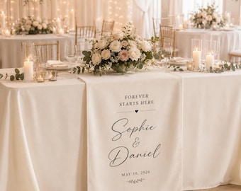 Puede incluir: Una mesa de recepción de bodas con un mantel crema y centros de mesa florales. Un camino de mesa muestra el texto "Forever Starts Here", los nombres "Sophie & Daniel" y la fecha "18 de mayo de 2026".