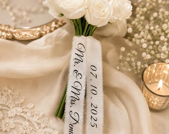 Puede incluir: Un ramo de rosas blancas con tallos verdes envuelto en dos cintas blancas. Una cinta presenta el texto impreso en negro Mr. & Mrs. Demir, y la segunda cinta muestra la fecha 07. 10. 2025 en una fuente serif negra a juego.