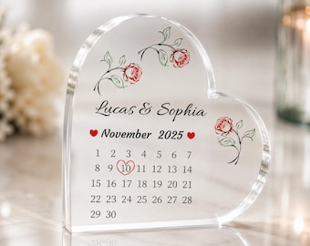 Puede incluir: Un calendario de acrílico transparente en forma de corazón con los nombres "Lucas & Sophia" y la fecha "Noviembre 2025." El número 10 está circulado. Rosas rojas y hojas verdes decoran la parte superior y lateral.