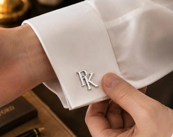 Puede incluir: Un gemelo de metal plateado con las letras P y K en una fuente con serifa, sujeto al puño de tela blanca de una camisa de manga larga. El gemelo presenta un acabado metálico pulido y está asegurado a través de los ojales de la camisa.