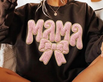 Puede incluir: Una sudadera marrón oscuro con la palabra "MAMA" en letras rosas, de estilo tejido. Una cinta rosa está debajo de la palabra "MAMA". Las letras y la cinta tienen un contorno dorado.