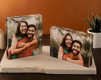 Puede incluir: Dos bloques de fotos rectangulares que muestran a una pareja abrazándose, colocados sobre una base de piedra de color claro. Las imágenes muestran a una mujer y un hombre sonriendo. Una planta suculenta en maceta está en el fondo.