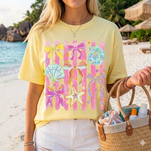 May include: Yellow t-shirt with a graphic of bows, seashells, and starfish. The design features pink vertical stripes and colorful bows. The shirt is worn with white shorts and a straw tote bag.