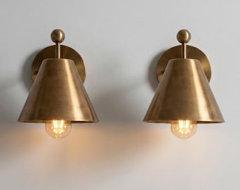 May include: A pair of wall-mounted sconces featuring a brushed brass finish. Each fixture consists of a circular backplate, a vertical stem topped with a small sphere, and a wide, conical shade housing a single glowing light bulb at the base.