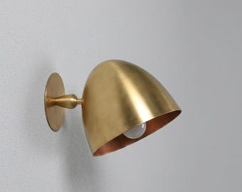 May include: A wall-mounted lamp featuring a dome-shaped shade with a brushed brass finish. The fixture includes a circular wall plate connected to the shade by a short, tapered arm, with a single white light bulb visible inside the metal housing.