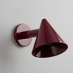 Puede incluir: Una lámpara de pared de color burdeos profundo con una base circular, un brazo cilíndrico corto y una pantalla cónica ancha. El aplique tiene un acabado liso y brillante, inclinado hacia abajo para dirigir la luz desde su interior.