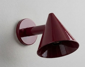 May include: A deep burgundy wall-mounted lamp featuring a circular base plate, a short cylindrical arm, and a wide, conical shade. The fixture has a smooth, glossy finish and is angled downward to direct light from the open, hollow interior.