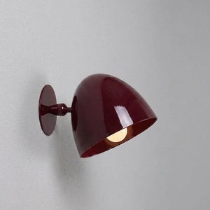 Puede incluir: Una lámpara de pared con una pantalla brillante en forma de cúpula de color burdeos intenso. El aplique incluye una placa base circular y un brazo corto a juego que se conecta al exterior redondeado y pulido, dejando ver una bombilla cálida.