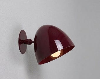 May include: A wall-mounted lamp featuring a glossy, deep burgundy, dome-shaped shade. The fixture includes a circular base plate and a short, matching arm that connects to the rounded, polished exterior, revealing a single warm light bulb inside.