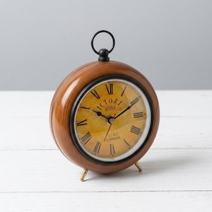 Puede incluir: Un reloj redondo de madera marrón con un asa negra y patas doradas. La esfera del reloj es amarilla con números romanos negros y las palabras "VICTORI STATION 1747 LONDON".
