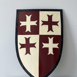 Op de afbeelding: Een schild in middeleeuwse stijl, met een zwarte rand en klinknageldetails. Het oppervlak van het schild is verdeeld in vier secties, afwisselend crème en bordeaux, elk met een wit kruis. Het schild heeft een licht gebogen vorm.
