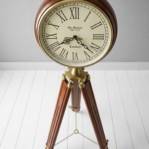 May include: A decorative clock with a cream face and black Roman numerals, mounted on a wooden tripod stand. The clock face features the text "Dbs Santos Estb. 1772 London". The clock has a dark brown wooden casing.