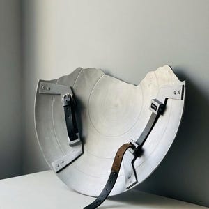 以下が含まれることがあります： 破損したエッジを持つ銀色のシールドで、黒い革のストラップと金属製のブラケットが付いています。 シールドは中央にへこみのある円形のデザインで、ニュートラルな背景に表示されています。