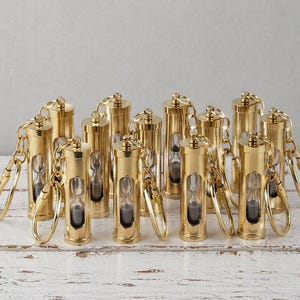 Könnte beinhalten: Eine Sammlung goldfarbener Sanduhr-Schlüsselanhänger. Jeder Schlüsselanhänger hat eine kleine Glas-Sanduhr, gefüllt mit dunklem Sand, in einem zylindrischen Metallrahmen. Die Schlüsselanhänger sind auf einer weißen Holzoberfläche angeordnet.