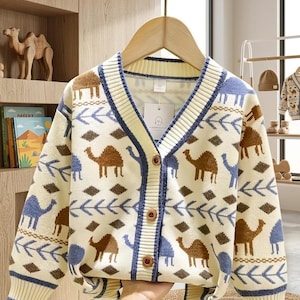 May include: Cream-colored cardigan with a camel pattern in brown and blue. The V-neck sweater has a blue trim and wooden buttons. The design includes diamond and arrow patterns. The tag reads "ZANANILO".