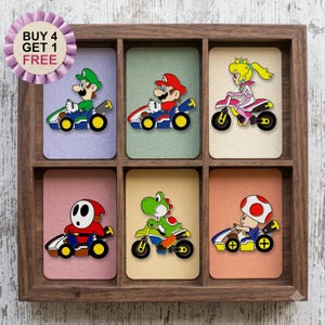Puede incluir: Una caja de madera con seis tarjetas rectangulares, cada una con un personaje diferente de la franquicia Mario conduciendo un kart o una motocicleta. Una insignia circular morada muestra el texto BUY 4 GET 1 FREE.