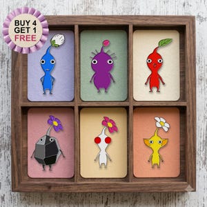 以下が含まれることがあります： 木製のディスプレイボックスに、個別のカードに描かれたカラフルなピクミンのキャラクターのピンバッジが6つ入っています。ピンバッジには、青、紫、赤、岩、白、黄色のキャラクターが含まれています。BUY 4 GET 1 FREEと書かれた紫色のバッジが添えられています。