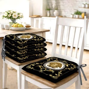 Peut inclure: Coussins de siège noirs avec un motif de tête de Méduse dorée et blanche. Les coussins carrés ont des volutes dorées et sont attachés à la chaise avec des rubans gris. Une pile de coussins est posée sur une table en bois. Dimensions : environ 40 cm x 40 cm.
