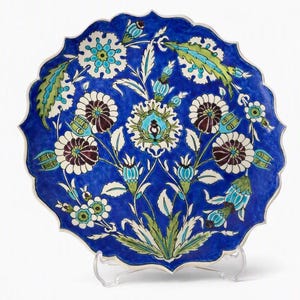 May include: A round ceramic plate with a scalloped edge, featuring a vibrant cobalt blue background decorated with intricate floral patterns in white, dark red, and shades of green. The design includes stylized blossoms, buds, and leaves.