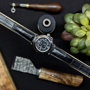 Può includere: Un orologio di lusso con cinturino in pelle nera e quadrante argentato. L'orologio è posto su uno sfondo nero con strumenti di orologeria e una piccola pianta succulenta. Il cinturino è lungo circa 23 cm.