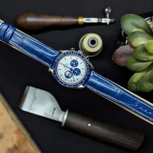 Peut inclure: Une montre de luxe avec un bracelet en cuir bleu et un cadran blanc avec des détails bleus. La montre est présentée sur une surface en bois avec des outils d'horlogerie et une petite plante. Le bracelet mesure environ 2 cm de large.