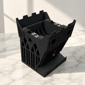 Peut inclure: Un accessoire de jeu imprimé en 3D noir en forme de château, doté de cadrans rotatifs intégrés pour suivre les statistiques de jeu. La base porte le texte en relief "War is Eternal" et le sommet présente des murs crénelés.