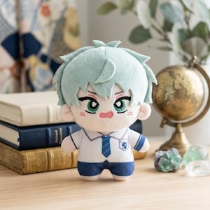May include: A plush doll with light blue hair and green eyes, wearing a white shirt with a blue tie and shorts. The doll has a small, embroidered face and is standing on a wooden surface. The doll is a collectible item.