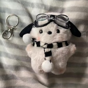 May include: A white dog plush keychain with black ears, wearing clear goggles and a black and white striped scarf. A silver keychain ring is next to the plush toy.