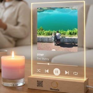 Op de afbeelding: Een verlichte acryl muziekdisplay met een foto van een stel bij een turquoise meer. De display toont de songtitel "DTMF" van Bad Bunny, met afspeelknoppen en een houten voet. Een brandende kaars en kleine potplanten staan ook op de tafel.