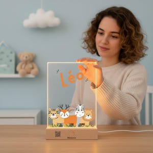 Può includere: Luce notturna a LED rettangolare con base in legno. La luce presenta un pannello trasparente con il nome "Léo" in arancione e animali dei cartoni animati. Gli animali includono una tigre, un cervo, una zebra e un leone. La base ha la scritta "Memoria".