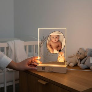 Può includere: Luce notturna personalizzata con foto di un bambino all'interno di una cornice circolare, con il nome "Theo" e un motivo a foglie. Una piccola luce a forma di volpe si trova sotto la foto. La base è in legno.