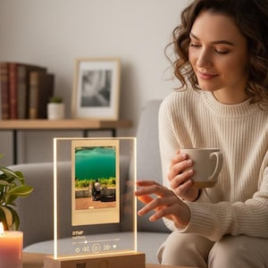 Può includere: Una cornice fotografica in acrilico trasparente con base in legno, illuminata con luce calda. La cornice mostra una foto di un paesaggio lacustre e controlli musicali. I testi "DTMF" e "Memoria" sono visibili. Una donna tiene una tazza.