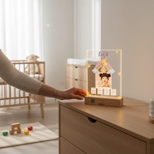 Può includere: Una luce notturna personalizzata in acrilico con base in legno. La luce presenta una foto di un bambino, un arcobaleno, una giraffa e il nome "Lola". La luce è su un comò in legno in una cameretta.