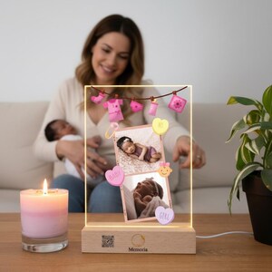 Può includere: Un espositore fotografico illuminato con due foto di neonati, dettagli rosa e viola e la scritta "Memoria" sulla base in legno. Sono visibili anche una candela accesa e una pianta.