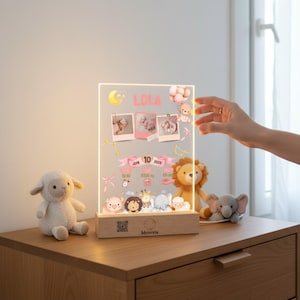 Può includere: Luce notturna personalizzata in acrilico con il nome "LOLA" in rosa, con foto del bambino e dettagli della nascita. Include una base in legno con illustrazioni di animali e un agnello e un leone di peluche.